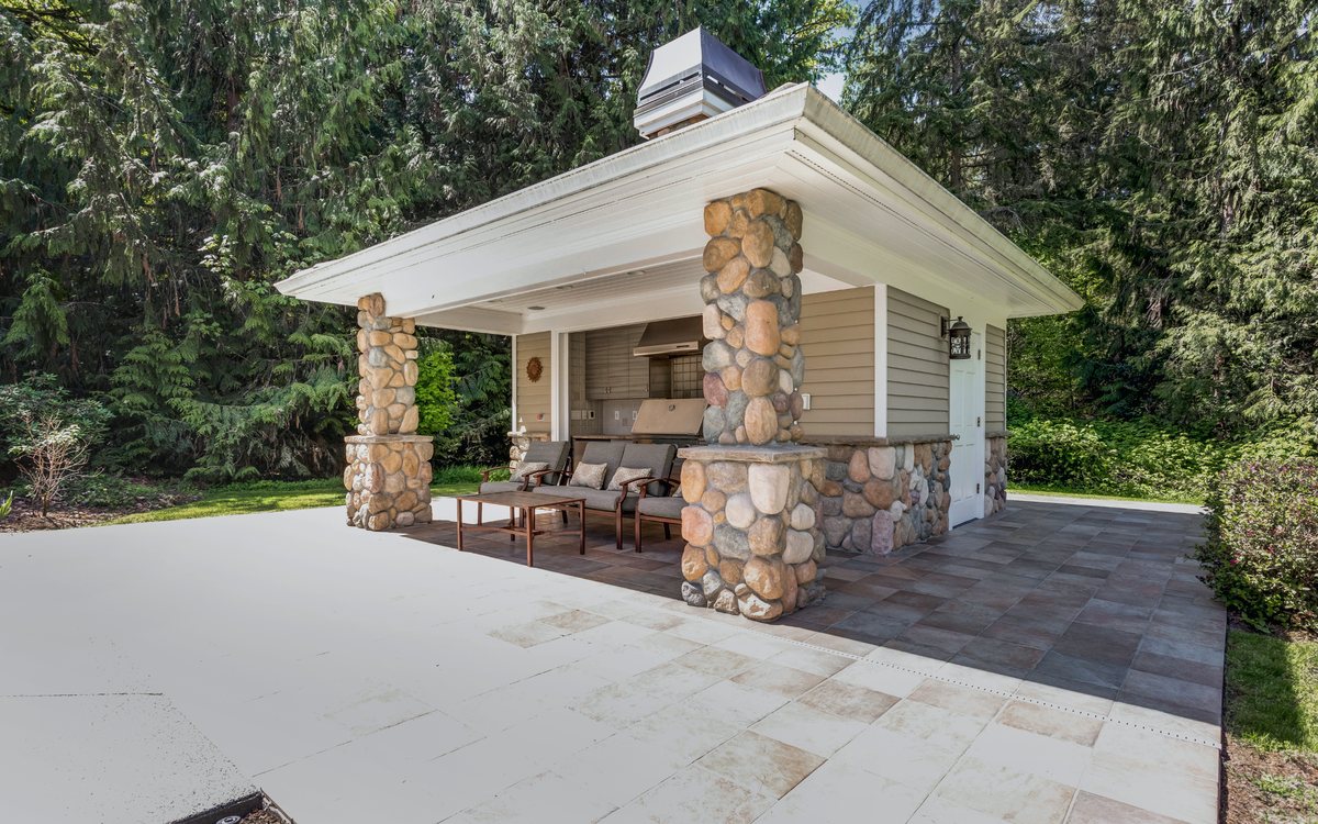 Outdoor living space with a stone clad kitchen and fireplace under a shingled roof, opening onto a paver patio