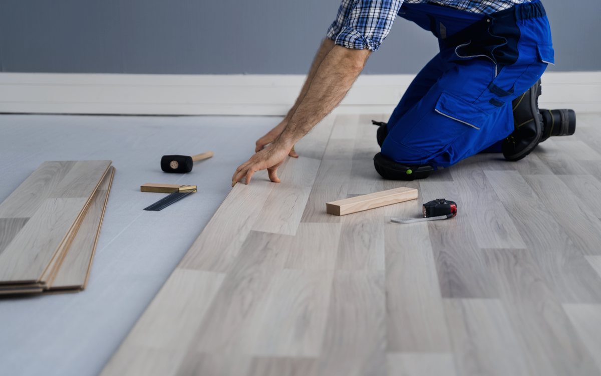 Installer fitting a plank of luxury vinyl flooring into place, with a tapping block, pull bar, and tape measure on the floor