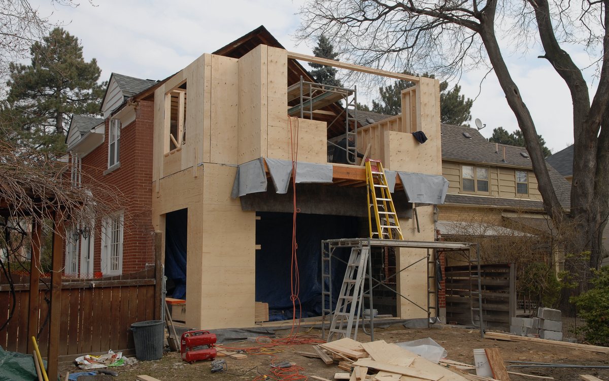Two story room addition under construction with new framing, sheathing, and scaffolding against the existing house