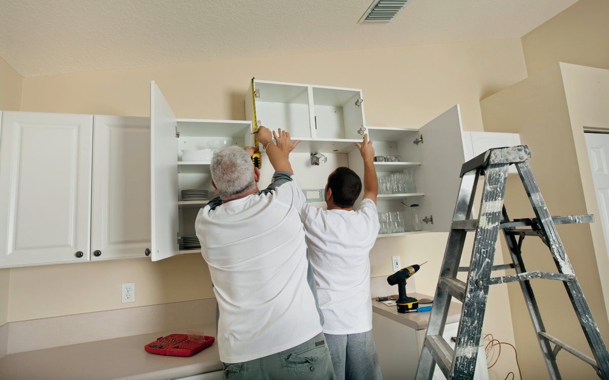 Two carpenters measuring and setting a white upper cabinet during a kitchen remodel, with a level and cordless drill on the counter