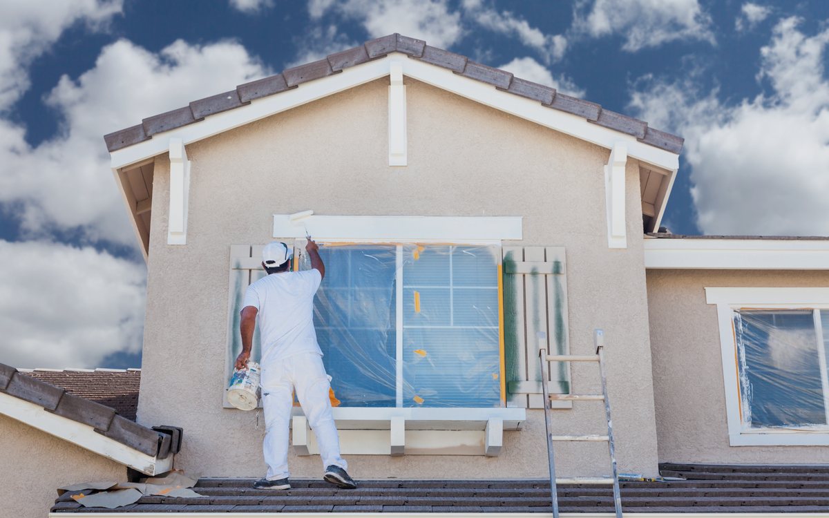 Painter cutting in around a window on a stucco exterior from a ladder, with the trim masked off