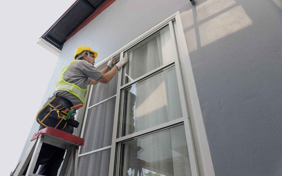 Installer setting a new exterior window into its opening from a ladder, checking the frame before fastening